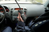 Zdjęcie kolorowe, przedstawiające policjanta znajdującego się w radiowozie, trzymającego radiotelefon.